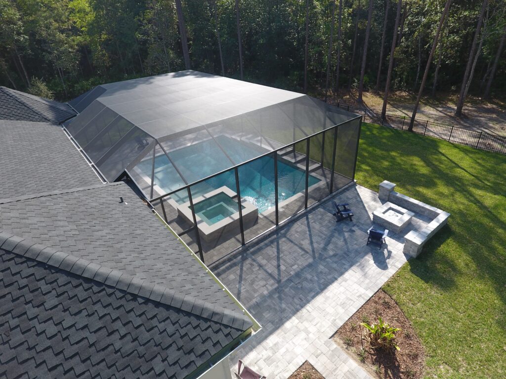 Aerial view of a modern screen enclosure surrounding a pool, featuring a spa, stone patio, and outdoor seating area in a lush backyard setting.