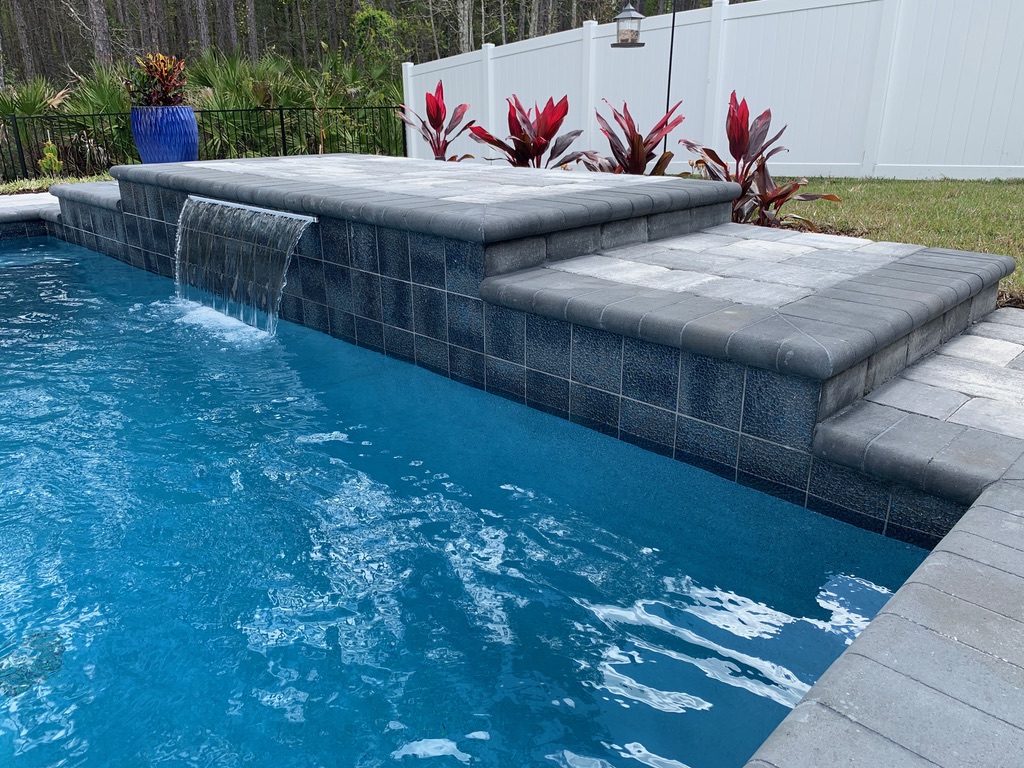 Waterfall feature cascading into a swimming pool, surrounded by paver stones and tropical plants, showcasing Breakwater Construction's design expertise.