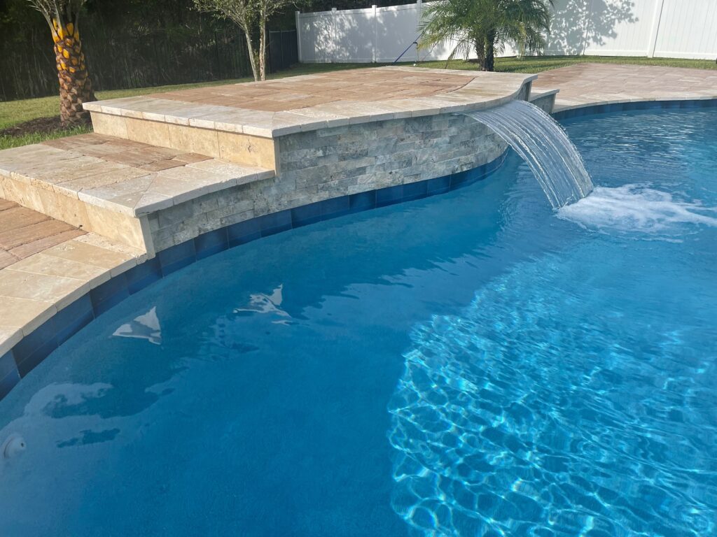 Waterfall feature cascading into a blue swimming pool, surrounded by stone and tile patio, showcasing Breakwater Construction's design expertise in water features.