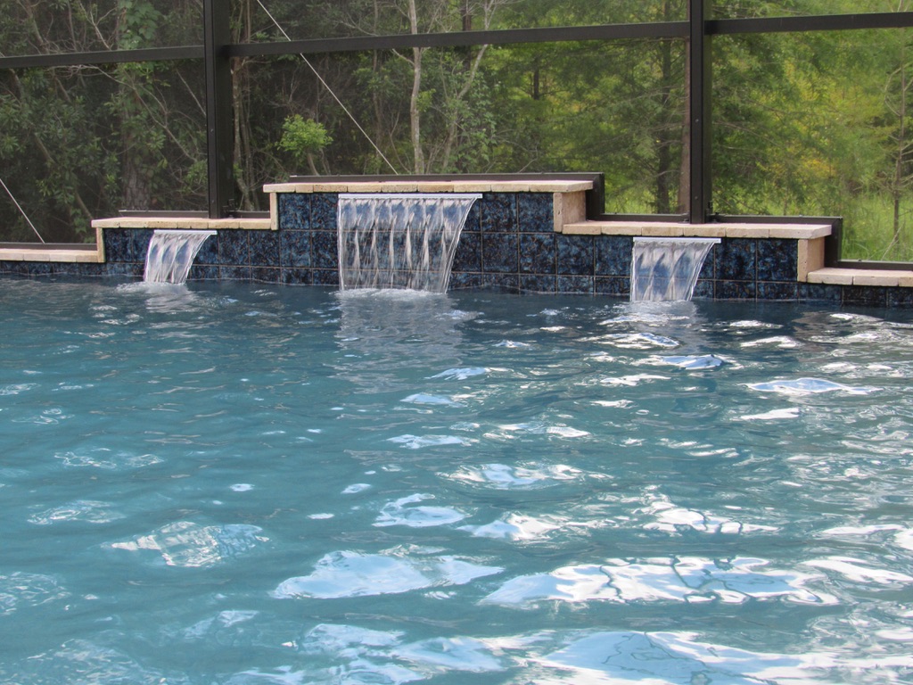 Waterfall feature cascading into a pool with blue tiles and natural greenery in the background, showcasing Breakwater Construction's design expertise.