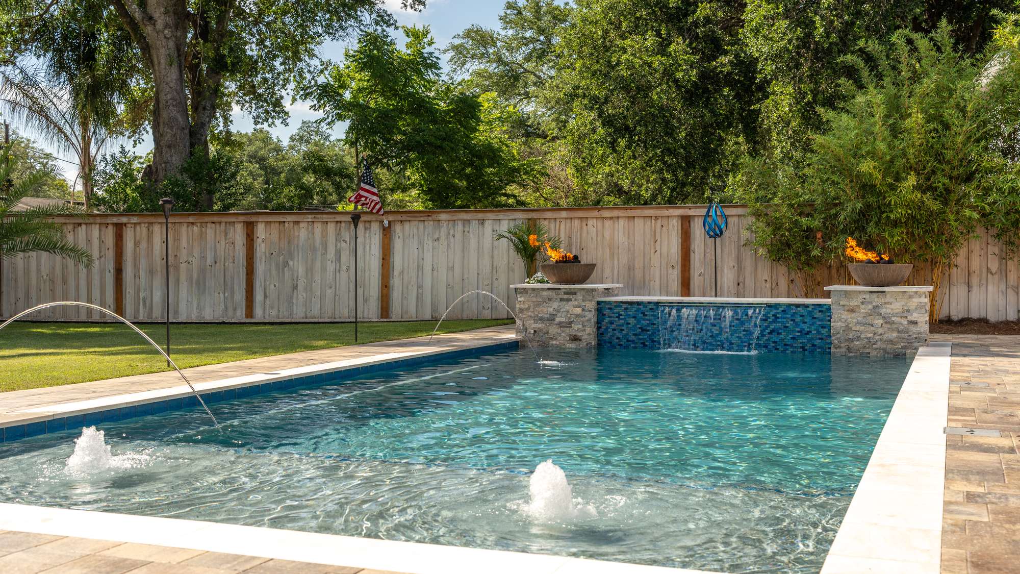 Wide view of a custom gunite pool with arcing deck jets, a blue glass tile spillway and two lit fire bowls, framed by a wood privacy fence and mature trees