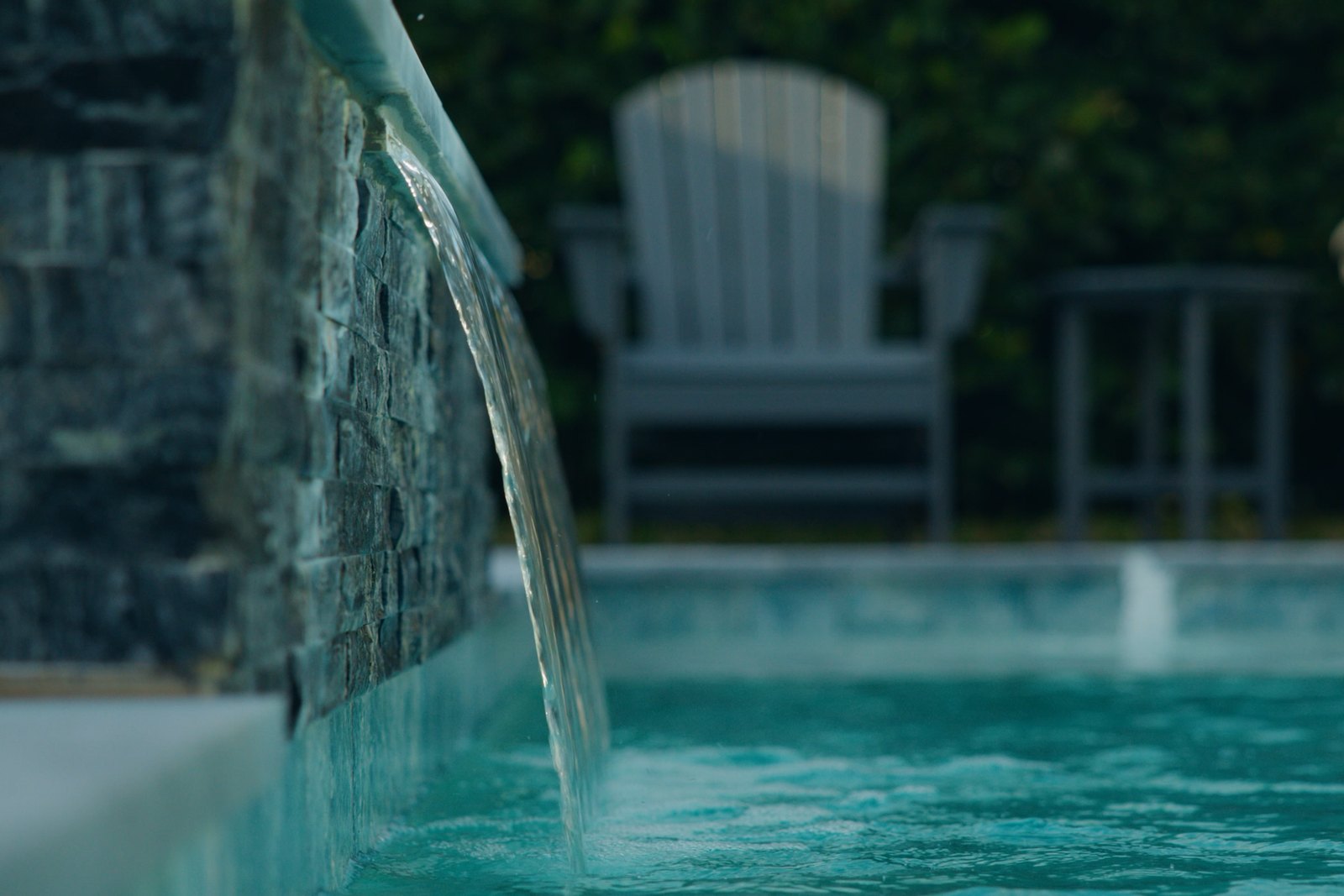 Water spilling from a raised stone water feature into a tiled pool beside finished decking and patio chairs