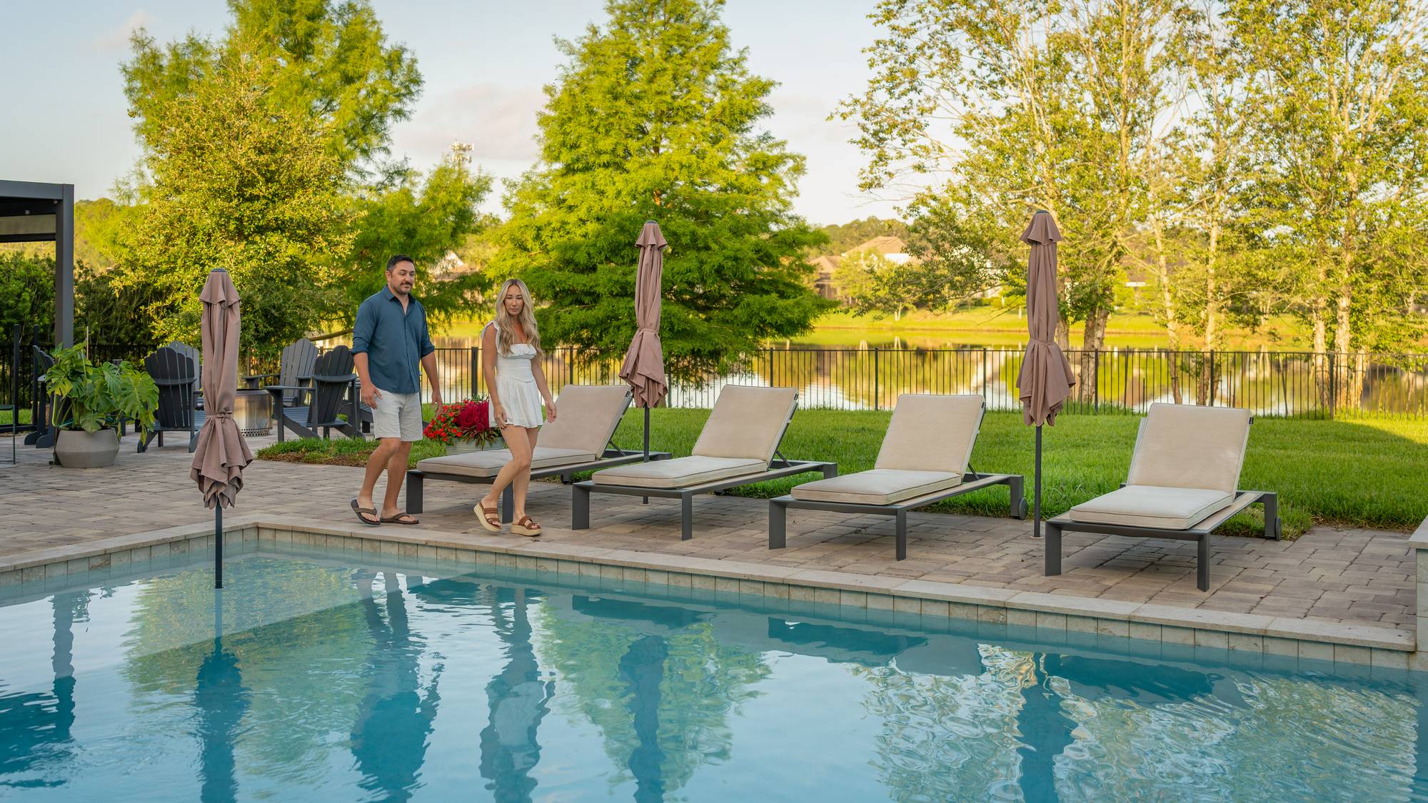 A couple walking along the paver deck beside a custom gunite pool at golden hour, with cushioned loungers, patio umbrellas and a pond behind them